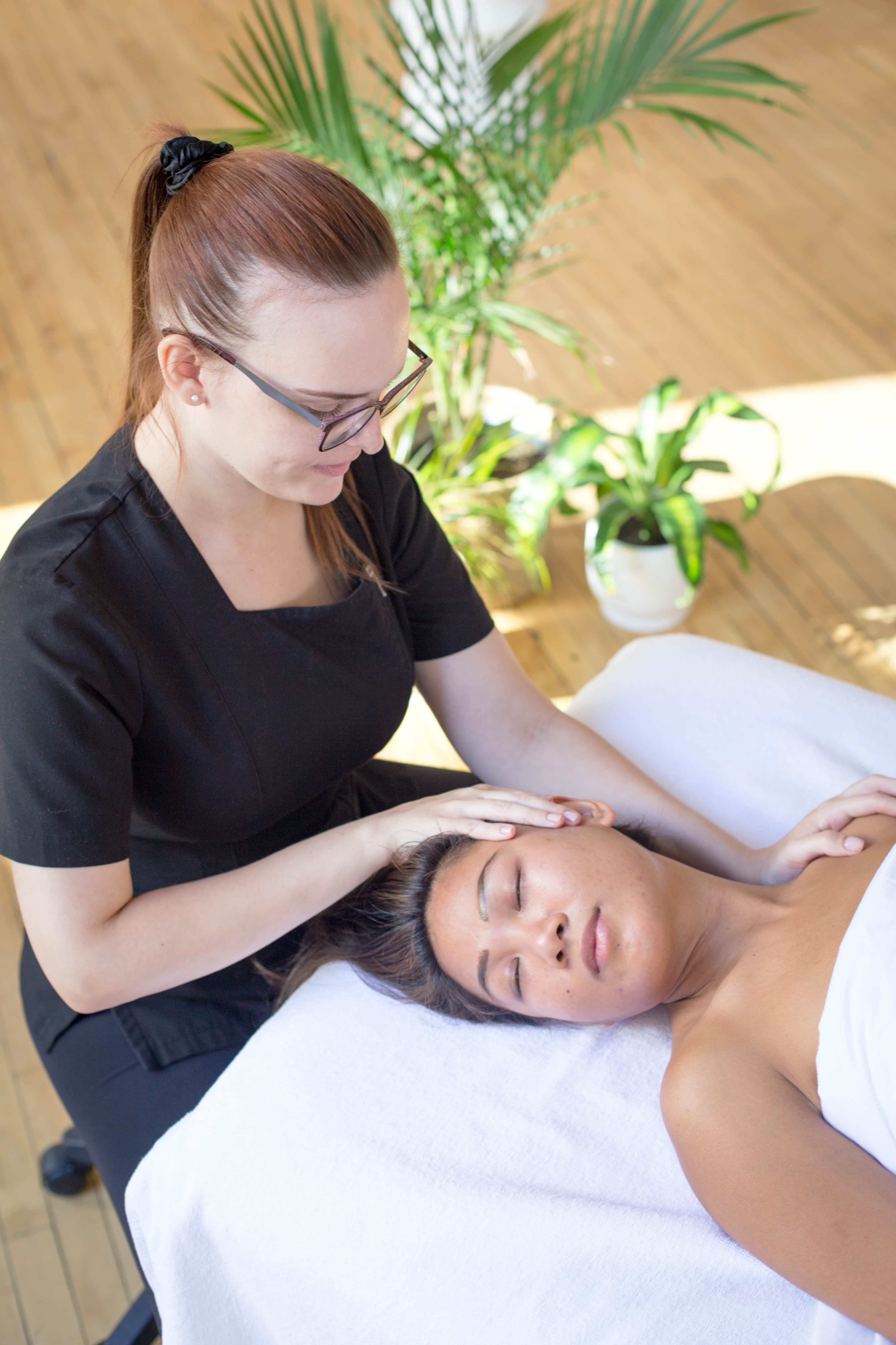 A woman lies on a massage table, face up under a cover. An esthetician massages her head and neck. The client looks relaxed.