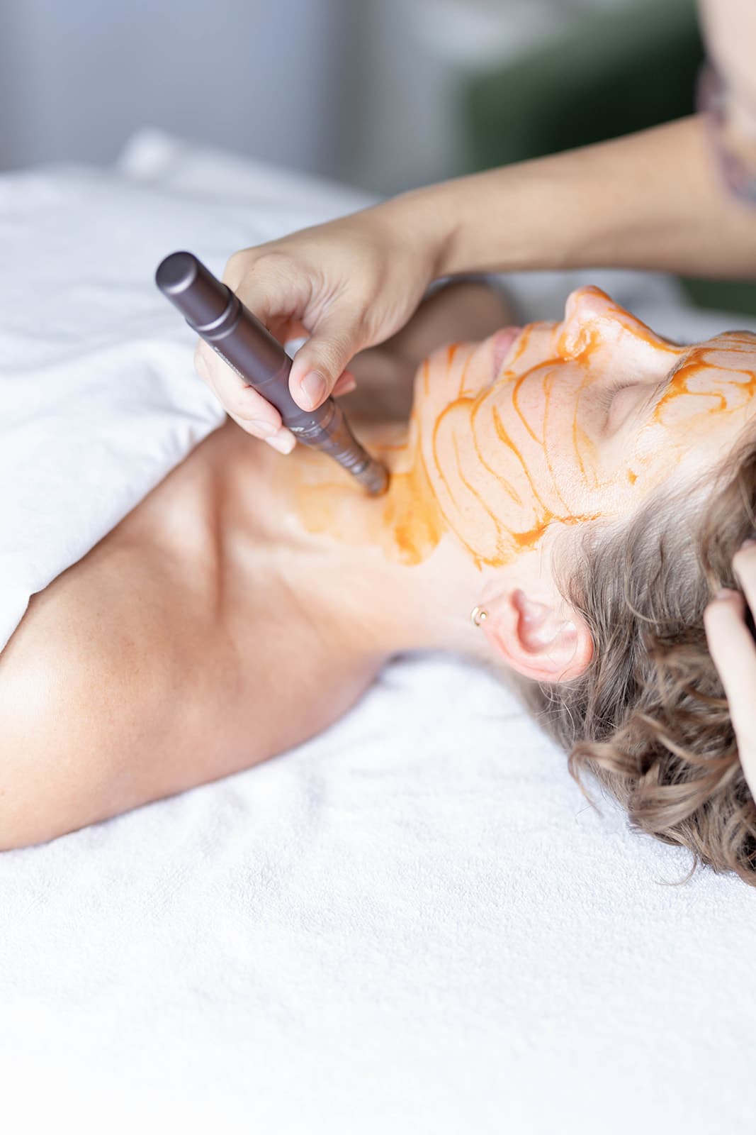 A client lays on her back with half her body exposed. An esthetician applies moisturizer to her client's leg